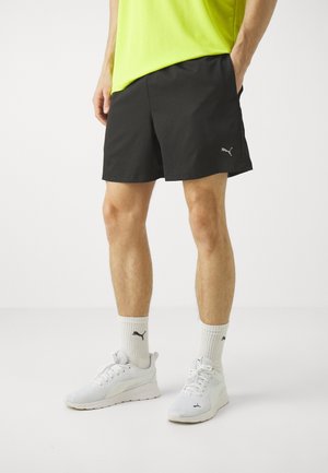 Mand iført sorte Puma-shorts, hvide Puma-strømper og hvide Puma-sneakers står mod en ensfarvet baggrund.