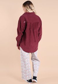 Chemise en velours côtelé bordeaux avec un col et un pli au dos ; associée à un pantalon à large jambe à imprimé floral et des baskets noires.