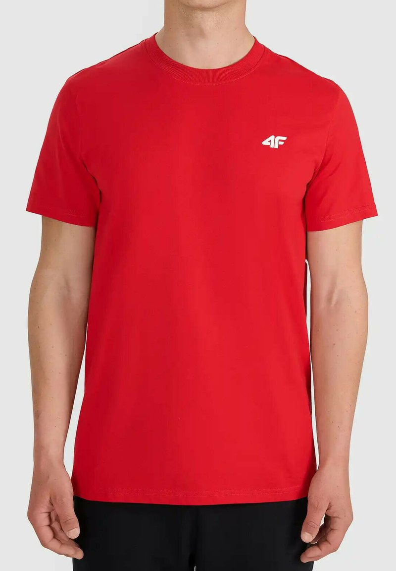 Homme portant un t-shirt rouge uni à manches courtes avec un petit logo blanc 4F sur la poitrine gauche, associé à un pantalon noir, debout devant un fond neutre.