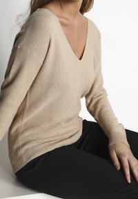 Beige stickad tröja med V-ringning och långa ärmar, med ribbad nederkant och muddar, bärs över svarta byxor. Mjuk textur, avslappnad passform.