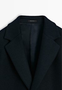 Dunkelblauer Wollmantel mit breiten Revers, sichtbarem schwarzen Futter und einem "Made in Portugal"- Etikett im Kragen.