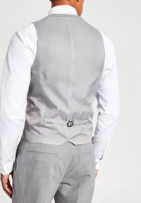 Graue Weste mit glatter Oberfläche, mit einem Schnallenversteller auf der Rückseite, über einem weißen Hemd getragen, kombiniert mit grauen Hosen.
