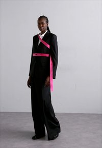 Costume noir avec pantalon large, chemise blanche et une ceinture rose vif sur la poitrine, avec un détail de bouton. Tissu lisse et design ajusté.