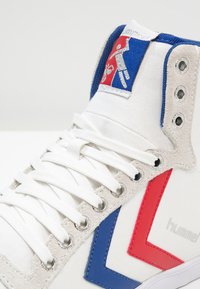 Sneaker înalt cu un upper din țesătură albă, accente din suede gri și detalii din cauciuc roșu și albastru. Include șireturi albe și ochiuri metalice.