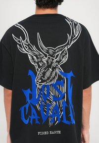 Svart oversized t-skjorte med et hvitt og blått grafisk motiv av en hjort med gevir og med bold tekst "Just Cavalli" og "Fired Earth" under.