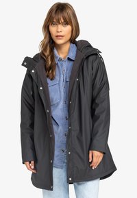 Zwarte waterdichte jas met een capuchon, voorzien van drukknoopsluiten, een gestructureerde antracietvoering en zijzakken. Gemaakt over een lichtblauw corduroy shirt.
