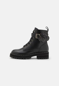 Botas de cuero negro hasta el tobillo con suela gruesa, herrajes dorados, diseño de cordones y un adorno floral en el costado.