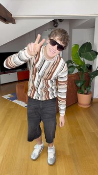 Suéter a rayas en tonos de marrón, blanco y negro; pantalones cortos de mezclilla negros; zapatillas plateadas; grandes gafas de sol negras; entorno interior.