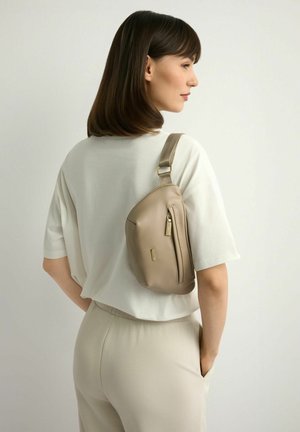 Kobieta ubrana w beżowy top z krótkim rękawem i spodnie, niosąca beżową skórzaną torebkę na ramię, stojąca na jednolitym tle.