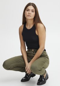 Zwarte mouwloze top, olijfgroene taps toelopende broek met knoopdetails en glanzende zwarte schoenen. Model hurkt met een neutrale uitdrukking.