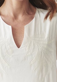 Haut en lin blanc avec un décolleté en V et de délicates broderies blanches de motifs en forme de feuilles de chaque côté de la zone de la poitrine.