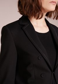 Černý blejzr s vroubkovaným klopem, zapínáním na dva knoflíky a bílými švy. Hladká tkanina a přiléhavý střih s ostrými švy.