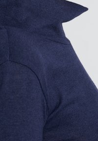 Mörkblå sweater med ett texturerat stickat mönster, med hög krage och en figurnära silhuett. Söm detaljer syns nära axlarna.