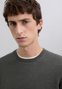 Grauer Pullover mit rundem Ausschnitt, gerippten Bündchen und einem dezenten Logo auf der linken Brust. Das Material wirkt weich und strukturiert.