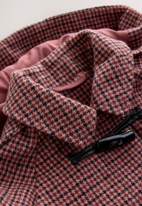 Tissu à motif pied-de-poule rose et noir avec une texture douce. Il présente un col et un bouton noir, avec une doublure rose clair visible à l'intérieur.