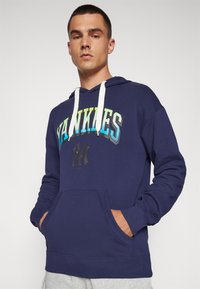 Navy plavi dukserica s grafikom "Yankees" u gradijent bojama, prednji džep i bijela špaga. Izrađena od mješavine pamuka s rebrastim manžetama.