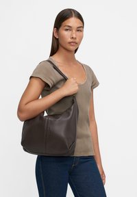 Borsa a spalla in pelle marrone con una superficie liscia, forma curvata e chiusura con zip; presentata con un modello che indossa un top aderente e jeans.