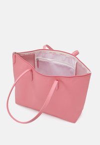 Sac fourre-tout rose en matériau synthétique lisse, avec des doubles poignées, une poche intérieure zippée et le logo Lacoste le long du bord intérieur.