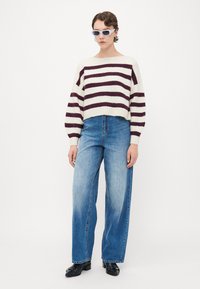 Person, der einen gestreiften weißen und lila Pullover, weite blaue Jeans, schwarze Schuhe und weiße Sonnenbrille trägt, steht vor einem einfarbigen Hintergrund.