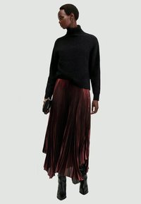 Maglione nero a collo alto e lunga gonna plissettata bordeaux. Tessuto liscio, parte superiore aderente, fondo svasato. Stivali neri fino al ginocchio e borsa piccola.