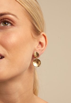 Mujer de piel clara y cabello rubio lleva un pendiente de doble disco dorado en su oreja izquierda, con un fondo beige.