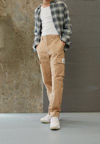 Avslappnad outfit med en grönrutig skjorta med knappar, beige cargobyxor med fickor och vita sneakers med blå detaljer.