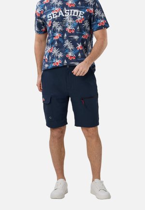 Mand iført marineblå cargoshorts og en blå blomster "SEASIDE" t-shirt med hvide sneakers, stående mod en hvid baggrund.