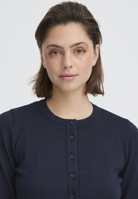 Cardigan bleu marine à boutons, fabriqué en tissu côtelé, doté d'un col rond et de six boutons sur le devant. Texture lisse, coupe ajustée.