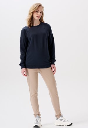 Marineblauwe sweatshirt met lange mouwen en ronde hals, gecombineerd met beige slim-fit joggingbroeken. Het model draagt witte en grijze sportschoenen.