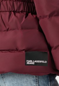 Detail blízko tmavě červené polstrované bundy s černou nálepkou s nápisem "KARL LAGERFELD JEANS" nad modrými džíny.