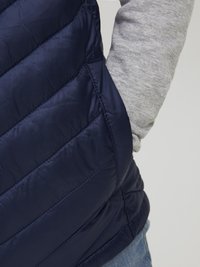 Námořnický prošívaný puffer vest s vertikálním šitím a hladkou texturou. Stažená látka u výřezů pro ruce, nošena přes šedé tričko s dlouhým rukávem.