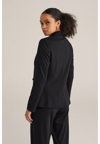 Crni sako s prilagođenim dizajnom, glatkom teksturom, zatvaranjem na jedan gumb i suptilnim detaljima šavova. Uparen s crnim hlačama.