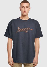 Tamsiai mėlynos spalvos trumpomis rankovėmis marškiniai, laisvo kirpimo, su oranžiniu "Rocawear" logotipu, atspausdintu priekyje. Pagaminti iš medvilnės, su apvali kaklo iškirpte.