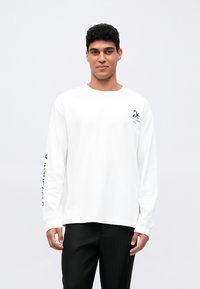 Weißes Langarm-T-Shirt mit rundem Halsausschnitt, das schwarzer grafischer Text entlang des linken Ärmels und ein kleines Logo auf der Brust zeigt.