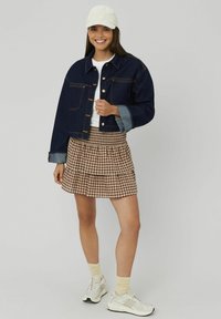 Jeune femme souriante, portant une casquette blanche, une veste en denim foncé, une chemise blanche, une jupe à carreaux bruns, des chaussettes crème et des baskets blanches.