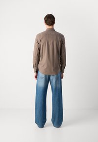 Camicia a maniche lunghe a righe verticali marroni, abbinate a jeans in denim chiaro a gamba larga. La camicia ha polsini con bottoni e un orlo curvo.