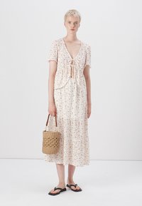 Robe maxi florale en tissu léger avec des manches courtes, un décolleté noué à l'avant, une jupe à étages et un sac en osier. Sandales plates noires.