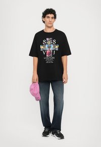 Black oversized t-shirt features a colorful graphic design with a cross pattern and "COUTURE" text; paired with faded blue jeans and a pink cap.