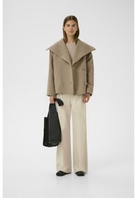 Camel wool blend jacket with a wide collar, double-breasted front, and two round buttons. Paired with cream wide-leg trousers and a black bag.
