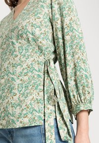Groene bloemenwrapblouse met lange, opgeblazen mouwen en een strikdetail op de taille. Gemaakt van lichte stof met een gestructureerd patroon.