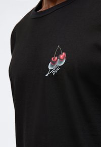 Tricou negru din bumbac, cu un design imprimat ce reprezintă două cireșe roșii și o ramură, cu numele brandului "Diesel" scris cu text alb dedesubt.