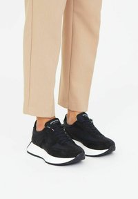 Zapatillas de ante negras con suela blanca, con cordones y logo en la lengüeta, combinadas con pantalones recortados de color beige claro.