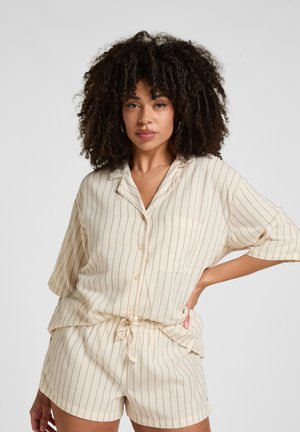 Frau mit lockigem Haar, die ein cremefarbenes, vertikal gestreiftes Kurzarmhemd und passende Shorts mit Kordelzug trägt, steht mit einer Hand in der Hüfte.