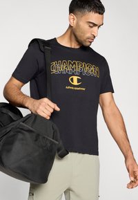 Μαύρο βαμβακερό μπλουζάκι με τη λέξη «CHAMPION» σε έντονα κίτρινα γράμματα, συνδυασμένο με μια μαύρη τσάντα τύπου duffel που διαθέτει δίχτυ τσέπης.