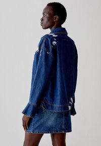 Giubbotto di denim con tessuto blu strappato, maniche lunghe, vestibilità oversize e tasca posteriore. Abbinato a una gonna corta di jeans coordinata.