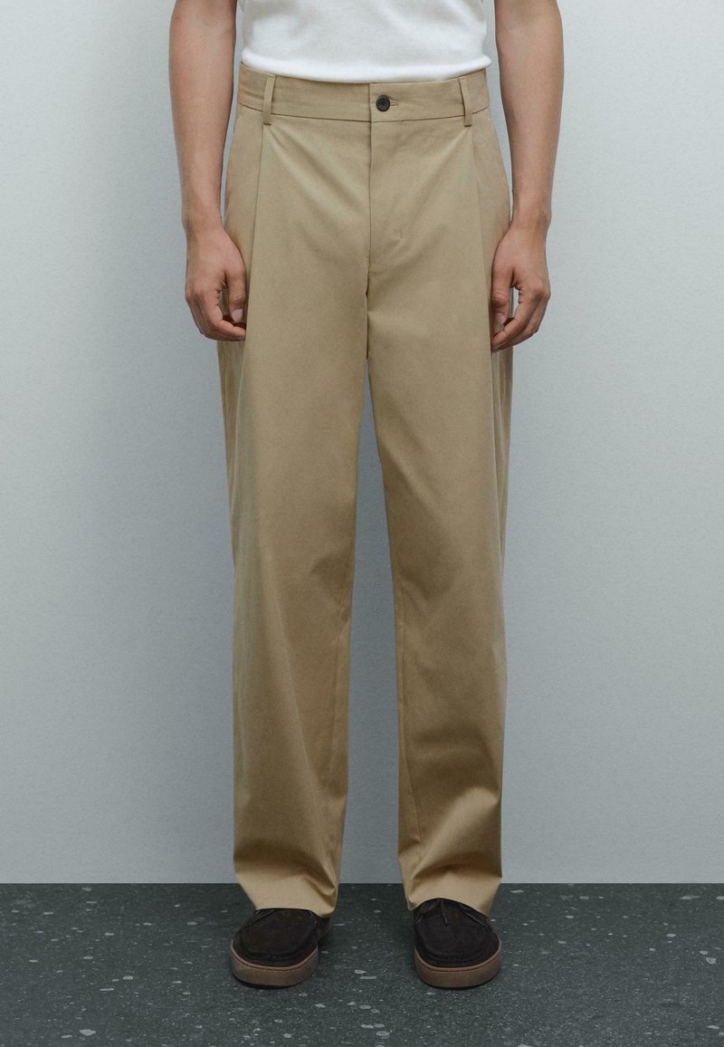 Beige, wide-leg trousers with pleats, made from a cotton blend. Includes a front button closure and side pockets. Worn with dark shoes.