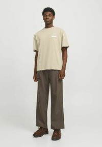 Beige bomulds t-shirt med løs pasform, korte ærmer og et lille hvidt logo. Kombinationen med mørkebrune vidde bukser og brune snørebåndssko.