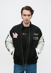 Νεαρός άνδρας με τα χέρια στις τσέπες φοράει μαύρο και λευκό μπλουζάκι τύπου varsity πάνω από μαύρο πουκάμισο Tommy Hilfiger και μπλε τζιν, σε απλό φόντο.