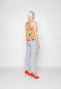 Top halter floral com padrões vibrantes em laranja e verde, combinado com jeans de denim azul claro e saltos altos vermelhos. Fundo simples.