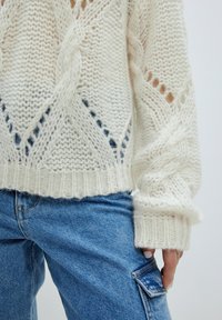 Weißer, gestrickter Pullover mit einem lässigen, strukturierten Design, offenen Mustern und einem gerippten Saum. Kombiniert mit hellblauen Jeans.